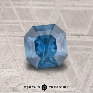 A 1.45-carat heated sapphire with an octagonal cut is displayed on a textured gray surface, with "EARTH'S TREASURY" printed below.