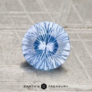 A 1.15-carat round, faceted Ceylon sapphire in light blue is displayed on a textured surface with the Earth’s Treasury logo at the bottom.