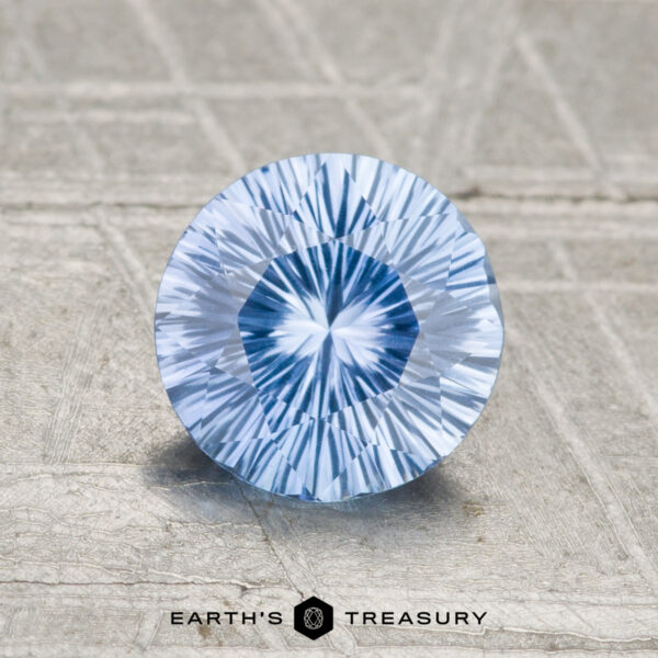 1.15-Carat Sky Blue Ceylon Sapphire A 1.15-carat round, faceted Ceylon sapphire in light blue is displayed on a textured surface with the Earth’s Treasury logo at the bottom.