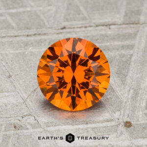 A 1.38-carat Spessartine Garnet, round and faceted with a vibrant orange hue, rests on a textured gray surface above the words "EARTH'S TREASURY.