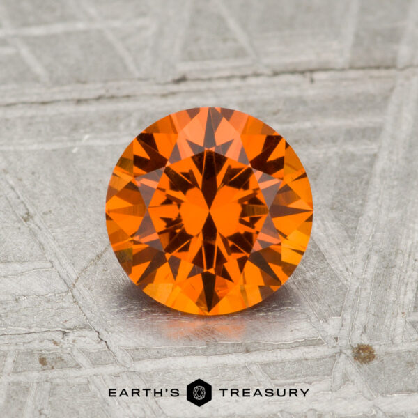 1.38-Carat Spessartine Garnet A 1.38-carat Spessartine Garnet, round and faceted with a vibrant orange hue, rests on a textured gray surface above the words "EARTH'S TREASURY.
