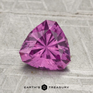 A 1.34-carat pastel pink pyrope garnet, faceted in a heart shape, rests on a textured gray surface with “EARTH'S TREASURY” displayed at the bottom.