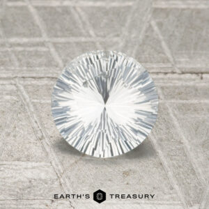 A 0.90-carat Ceylon Sapphire, round brilliant-cut and faceted, is displayed upright on a textured gray surface with the "EARTH'S TREASURY" logo and text at the bottom.