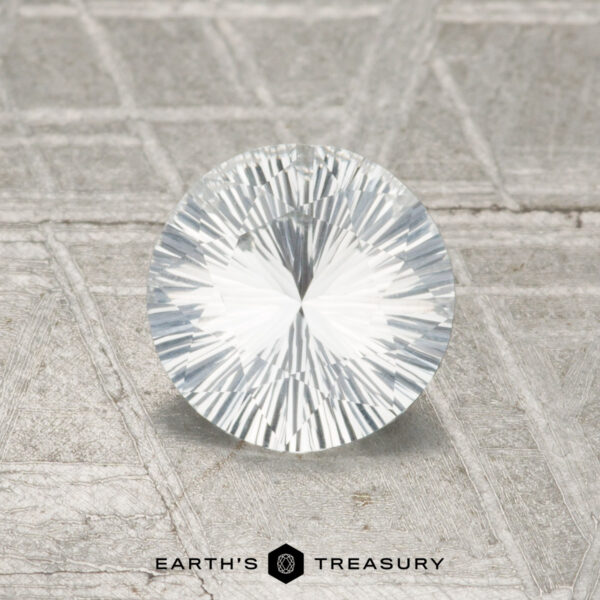 0.90-Carat Colorless Ceylon Sapphire A 0.90-carat Ceylon Sapphire, round brilliant-cut and faceted, is displayed upright on a textured gray surface with the "EARTH'S TREASURY" logo and text at the bottom.