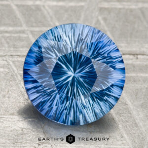 A 2.99-carat round Ceylon sapphire (heated), blue with intricate internal patterns, is displayed on a textured gray surface. "Earth's Treasury" appears as logo text at the bottom.