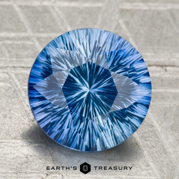 A 2.99-carat round Ceylon sapphire (heated), blue with intricate internal patterns, is displayed on a textured gray surface. "Earth's Treasury" appears as logo text at the bottom.