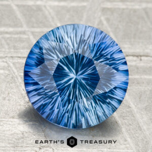 A 2.99-carat round Ceylon sapphire (heated), blue with intricate internal patterns, is displayed on a textured gray surface. "Earth's Treasury" appears as logo text at the bottom.