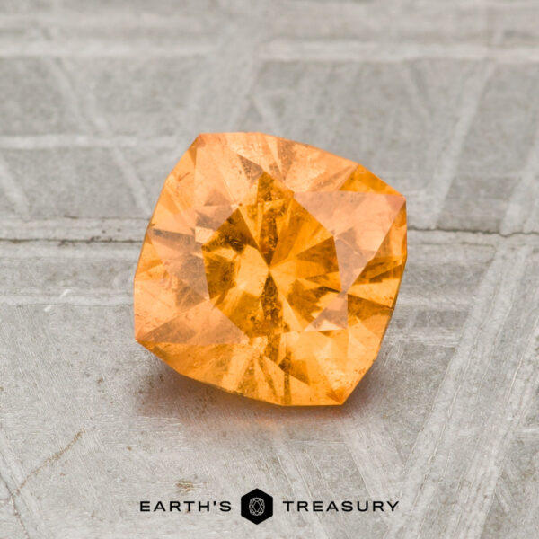 1.35-Carat Spessartine Garnet A 1.35-carat spessartine garnet, faceted in a vibrant orange-yellow hue, rests on a textured metallic surface with "EARTH'S TREASURY" displayed at the bottom.