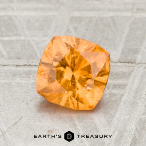 A 1.35-Carat Spessartine Garnet with a faceted cut and light orange hue rests on a textured gray surface, with the text "Earth's Treasury" displayed below.