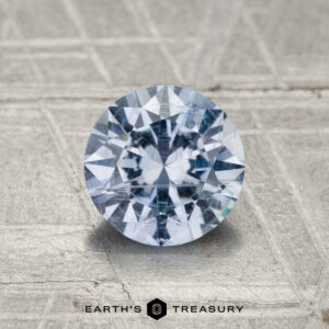 A 1.30-carat round brilliant-cut Montana Sapphire is displayed on a textured metallic surface, with "Earth's Treasury" shown at the bottom.