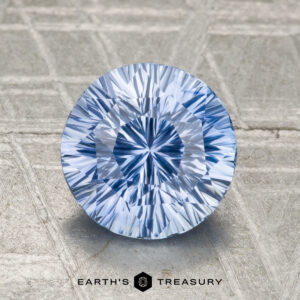 A round, faceted 2.01-carat heated Ceylon sapphire with a light blue hue stands upright on a textured surface, displayed above the text "EARTH'S TREASURY.