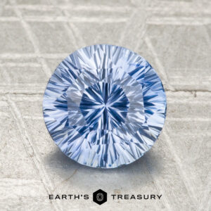 A round, faceted 2.01-carat heated Ceylon sapphire with a light blue hue stands upright on a textured surface, displayed above the text "EARTH'S TREASURY.