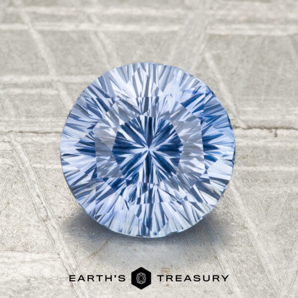 A round, faceted 2.01-carat heated Ceylon sapphire with a light blue hue stands upright on a textured surface, displayed above the text "EARTH'S TREASURY.