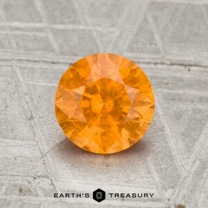 A 1.32-carat Spessartine Garnet, round brilliant-cut, is displayed on a textured grey surface with "EARTH'S TREASURY" at the bottom.