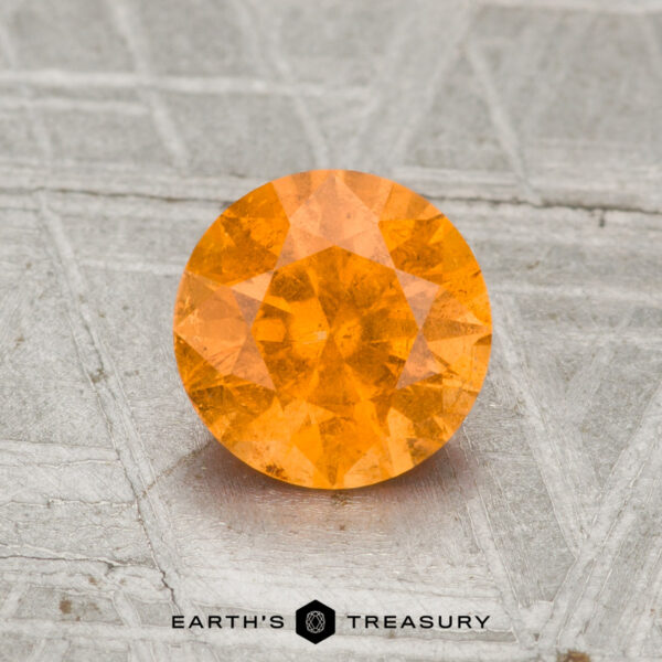 1.32-Carat Spessartine Garnet A 1.32-carat Spessartine Garnet, round brilliant-cut, is displayed on a textured grey surface with "EARTH'S TREASURY" at the bottom.