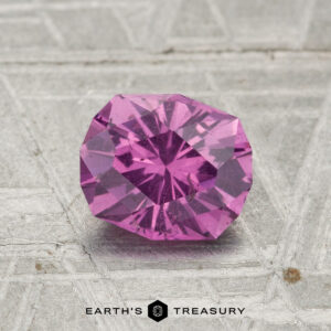 A 1.19-carat pastel pyrope garnet gemstone rests on a textured gray surface with “EARTH’S TREASURY” and a hexagonal logo below.