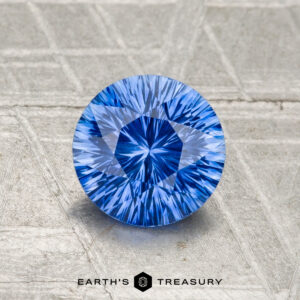 A 1.15-carat, round brilliant-cut Ceylon sapphire (heated) is showcased on a textured, light-colored surface with "EARTH'S TREASURY" at the bottom.