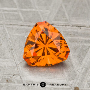 A 1.30-carat spessartine garnet, faceted in a triangular shape and vibrant orange, rests on a textured gray surface with "EARTH'S TREASURY" displayed at the bottom.