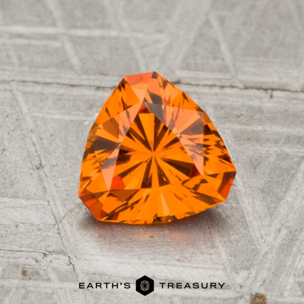 1.30-Carat Spessartine Garnet A 1.30-carat spessartine garnet, faceted in a triangular shape and vibrant orange, rests on a textured gray surface with "EARTH'S TREASURY" displayed at the bottom.