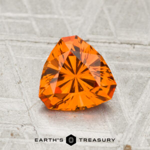 A 1.30-carat spessartine garnet, faceted in a triangular shape and vibrant orange, rests on a textured gray surface with "EARTH'S TREASURY" displayed at the bottom.