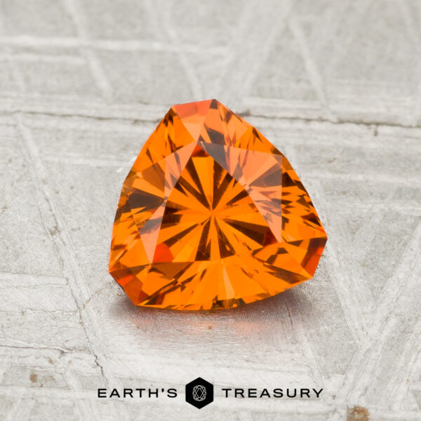 1.30-Carat Spessartine Garnet A 1.30-carat spessartine garnet, faceted in a triangular shape and vibrant orange, rests on a textured gray surface with "EARTH'S TREASURY" displayed at the bottom.