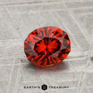 A 1.28-carat spessartine garnet, round and faceted, is shown on a textured gray surface with "EARTH'S TREASURY" at the bottom.