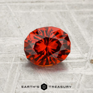 A 1.28-carat spessartine garnet, round and faceted, is shown on a textured gray surface with "EARTH'S TREASURY" at the bottom.