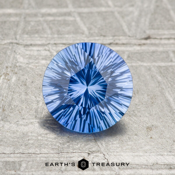 1.07-Carat Ceylon Sapphire (Heated) A 1.07-carat heated Ceylon sapphire, round and faceted with a starburst pattern, rests on a textured gray surface. The "EARTH'S TREASURY" logo appears at the bottom.