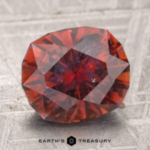 A 4.18-carat heated Montana sapphire, deep red with visible inclusions, rests on a textured metallic surface. "EARTH'S TREASURY" appears at the bottom.
