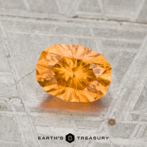 A 1.21-carat spessartine garnet with an oval cut rests on a textured gray surface, with "Earth's Treasury" displayed at the bottom of the image.