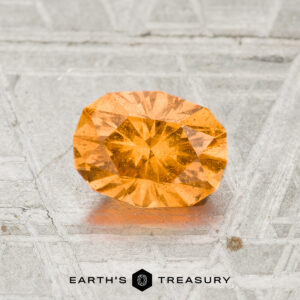 A 1.21-carat spessartine garnet with an oval cut rests on a textured gray surface, with "Earth's Treasury" displayed at the bottom of the image.