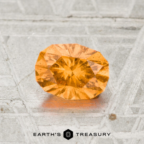 1.21-Carat Spessartine Garnet A 1.21-carat spessartine garnet with an oval cut rests on a textured gray surface, with "Earth's Treasury" displayed at the bottom of the image.
