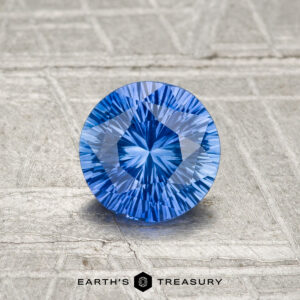 A 0.90-carat heated Ceylon sapphire, round and brilliant-cut, rests on a textured gray surface above the words "EARTH'S TREASURY" and a hexagonal logo.
