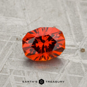 A 1.17-carat Spessartine garnet, expertly faceted in an oval cut, rests on a textured grey surface with "Earth's Treasury" displayed below.