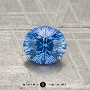 A 0.93-carat Ceylon sapphire, cut in a round brilliant style, rests on a textured metallic surface with "Earth's Treasury" displayed at the bottom.