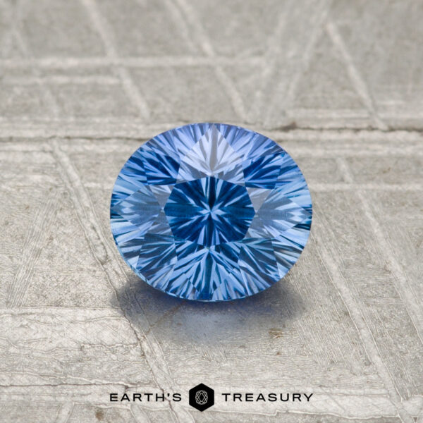 0.93-Carat Cornflower Blue Ceylon Sapphire A 0.93-carat Ceylon sapphire, cut in a round brilliant style, rests on a textured metallic surface with "Earth's Treasury" displayed at the bottom.
