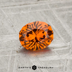 A 0.97-carat Spessartine garnet with vibrant orange facets rests on a textured gray surface, featuring the "EARTH'S TREASURY" logo at the bottom.