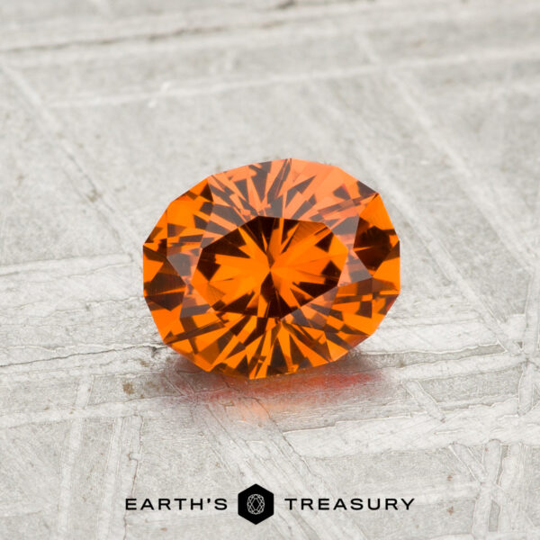 0.97-Carat Spessartine Garnet A 0.97-carat Spessartine garnet with vibrant orange facets rests on a textured gray surface, featuring the "EARTH'S TREASURY" logo at the bottom.
