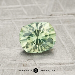 A 1.06-carat Montana sapphire, faceted in light green, is showcased on a textured gray surface with "EARTH'S TREASURY" at the bottom.