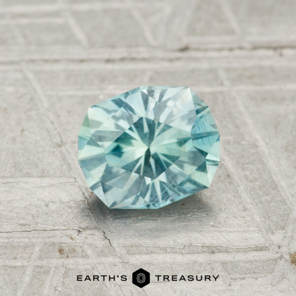 0.99-Carat Montana Sapphire A round, faceted 0.99-carat Montana Sapphire in light blue rests on a textured gray surface, with "EARTH'S TREASURY" text at the image’s bottom.
