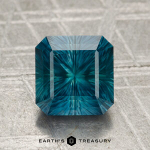A 2.24-carat heated Montana sapphire, emerald-cut in teal-blue with a radiant internal pattern, is shown on a textured gray surface with the "Earth's Treasury" logo below.