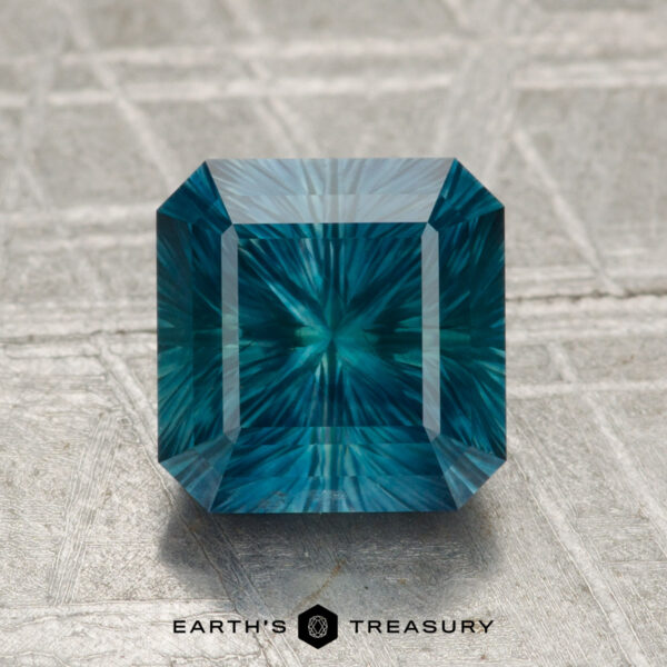 2.24-Carat Montana Sapphire (Heated) A 2.24-carat heated Montana sapphire, emerald-cut in teal-blue with a radiant internal pattern, is shown on a textured gray surface with the "Earth's Treasury" logo below.