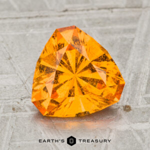 A 2.45-carat, faceted spessartine garnet with a vibrant orange hue is shown on a textured gray surface, featuring the text "EARTH'S TREASURY" at the bottom.