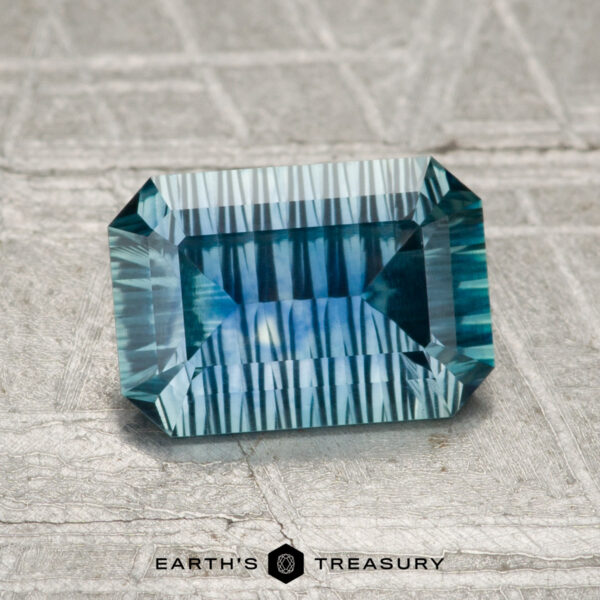 1.35-Carat Montana Sapphire (Heated) A rectangular, blue-green 1.35-carat Montana Sapphire (Heated) is displayed on a textured gray surface with "EARTH'S TREASURY" below.