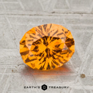 A 2.19-carat, faceted oval Spessartine Garnet with a vivid orange hue rests on a textured gray surface, with the text "EARTH'S TREASURY" displayed below.