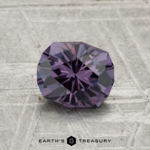 A faceted, round 1.21-carat color-change garnet is displayed on a textured gray surface with the text "EARTH'S TREASURY" at the bottom.