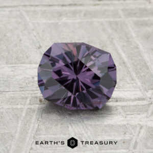 A faceted, round 1.21-carat color-change garnet is displayed on a textured gray surface with the text "EARTH'S TREASURY" at the bottom.