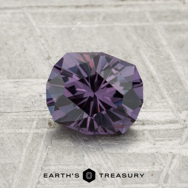 A faceted, round 1.21-carat color-change garnet is displayed on a textured gray surface with the text "EARTH'S TREASURY" at the bottom.