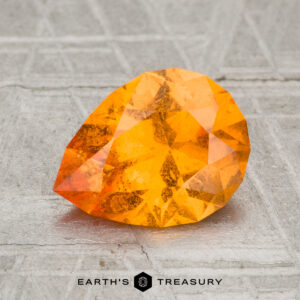 A faceted, pear-shaped 2.15-carat Spessartine Garnet is displayed on a textured gray surface with the "Earth's Treasury" logo beneath it.