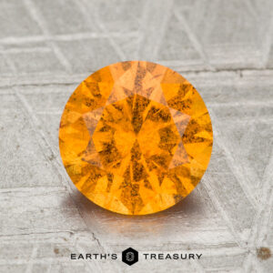 A 2.10-carat round, faceted Spessartine Garnet gemstone with an orange hue is displayed on a textured gray surface, with the text "Earth's Treasury" underneath.